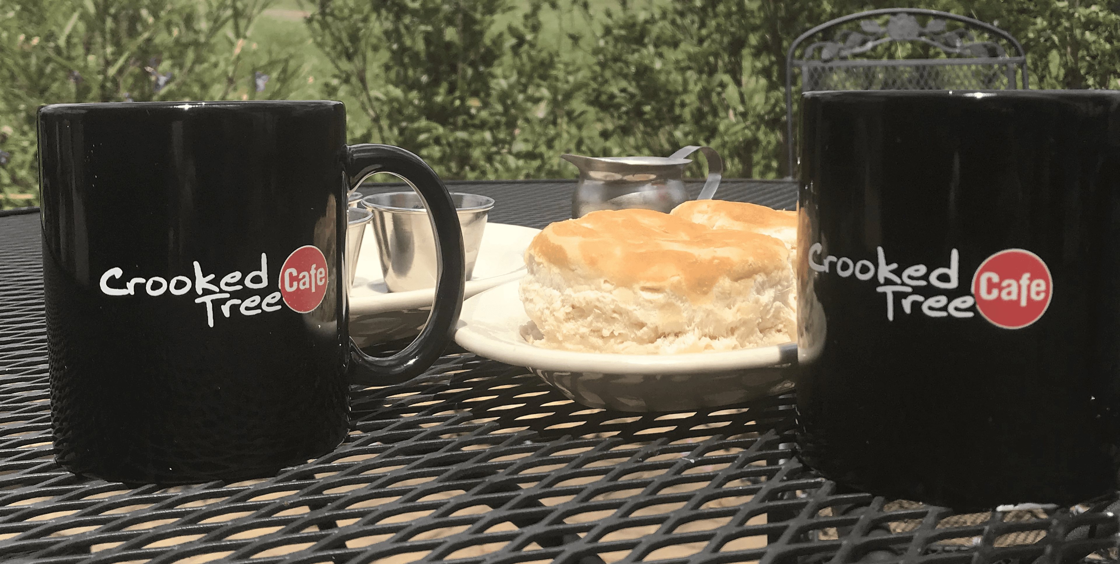 Two coffee mugs and a plate with a sandwich on a metal table outside.